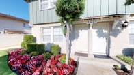 This single-story property presents an inviting streetscape defined by thoughtfully maintained landscaping and classic exterior design. The front yard features a well-kept lawn bordered by dark mulched garden beds filled with a variety of flowering plants, ornamental shrubs, and mature trees, including a prominent palm that adds a distinct coastal character to the setting. The neighborhood context suggests a sunny, temperate climate, consistent with Southern California living.

The building's exterior showcases a combination of brick and board-and-batten siding in a muted sage green, giving it a clean, traditional aesthetic. The entrance area is neatly appointed with a covered entry portico, a classic lantern-style wall light, and dual unit doors flanked by trimmed hedges. Vibrant flower beds featuring red, pink, and yellow blooms add a lively, colorful accent directly in front of the entrance.

The surrounding streetscape is lined with mature trees and well-maintained lawns, contributing to a calm, established neighborhood atmosphere. Overall, the property conveys a sense of careful upkeep, outdoor charm, and comfortable curb appeal suited to a pleasant residential environment.