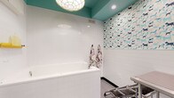 Located at 101–165 E Louisiana St in Indianapolis, this single-floor pet grooming facility is thoughtfully designed to provide a functional yet welcoming environment for both pets and their owners. The space centers around a dedicated grooming and bathing area outfitted with large, built-in white bathing tubs equipped with handheld spray attachments, making the bathing process efficient and comfortable. A stainless steel grooming table with an adjustable arm and professional-grade dryer adds to the facility's well-equipped setup. The interior design strikes a balance between clinical cleanliness and playful charm. White subway tile lines the lower walls, while a vibrant dog-patterned wallcovering adorns the upper portions, featuring illustrated dogs in shades of blue, teal, and navy. The teal-painted ceiling adds a bold pop of color overhead, complemented by a decorative pendant light fixture that gives the space personality. Dark charcoal doors with glass sidelights provide visual separation between zones while maintaining an open, airy feel. Whimsical animal-shaped wall hooks in yellow, green, white, and black serve as both functional accessories and decorative accents. Durable, easy-to-clean flooring throughout underscores the facility's practical design. Overall, the space combines professional grooming functionality with a lively, pet-friendly aesthetic.