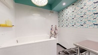 Located at 101–165 E Louisiana St in Indianapolis, this single-floor pet grooming facility is thoughtfully designed to provide a functional yet welcoming environment for both pets and their owners. The space centers around a dedicated grooming and bathing area outfitted with large, built-in white bathing tubs equipped with handheld spray attachments, making the bathing process efficient and comfortable. A stainless steel grooming table with an adjustable arm and professional-grade dryer adds to the facility's well-equipped setup. The interior design strikes a balance between clinical cleanliness and playful charm. White subway tile lines the lower walls, while a vibrant dog-patterned wallcovering adorns the upper portions, featuring illustrated dogs in shades of blue, teal, and navy. The teal-painted ceiling adds a bold pop of color overhead, complemented by a decorative pendant light fixture that gives the space personality. Dark charcoal doors with glass sidelights provide visual separation between zones while maintaining an open, airy feel. Whimsical animal-shaped wall hooks in yellow, green, white, and black serve as both functional accessories and decorative accents. Durable, easy-to-clean flooring throughout underscores the facility's practical design. Overall, the space combines professional grooming functionality with a lively, pet-friendly aesthetic.