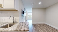 This efficiently designed single-floor unit offers a thoughtful layout across multiple living areas, a kitchen, a full bathroom, and a closet. Light-toned wood-look flooring runs consistently throughout the space, contributing to a clean, cohesive aesthetic.

The kitchen is appointed with white shaker-style cabinetry featuring gold hardware, striking granite countertops with bold veining, a classic white subway tile backsplash, and stainless steel appliances including a refrigerator, range, and over-the-range microwave. The galley-style layout maximizes functionality within a compact footprint.

The living areas are bright and open, with neutral warm-beige walls, recessed lighting, and a wall-mounted flat-screen television. Large windows with horizontal blinds allow generous natural light to fill the space, offering views of surrounding greenery.

The bedroom areas include closet storage with built-in hanging rods. One area also features an in-unit washer, adding everyday convenience. The bathroom is finished with a marble-look granite vanity countertop, an undermount sink, and a chrome faucet, maintaining the upscale material palette found throughout.

Overall, the unit presents a modern, polished interior that makes efficient and stylish use of its space.
