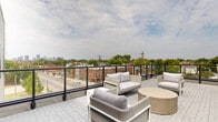 Located at 1179 E Main St in Columbus, Ohio's dynamic 43205 corridor, this rooftop terrace offers an impressive open-air retreat with sweeping views of the Columbus city skyline and the surrounding neighborhood. The terrace is finished with a sophisticated patterned paver deck that extends generously across the entire outdoor level, providing ample space for relaxation and social gatherings.

The space is thoughtfully divided into distinct lounge areas, each furnished with contemporary wicker and upholstered outdoor seating, including sofas, armchairs, and round drum-style side tables in neutral tones. One seating area is anchored near a sleek stone accent wall with a sliding glass door entry, creating a semi-sheltered nook ideal for more intimate gatherings. The perimeter of the terrace features a clean-lined black metal railing system, allowing unobstructed panoramic views of the Columbus skyline to the west, as well as the tree-lined streetscape below along E Main St.

The modern architectural exterior incorporates horizontal lap siding in a warm gray tone alongside light stone cladding, lending a polished, contemporary aesthetic to the rooftop structure. The open-sky setting floods the terrace with natural light throughout the day, making it a versatile space well-suited for both daytime lounging and evening gatherings in the heart of Columbus.