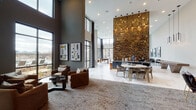 Located in Frisco, TX, this contemporary community clubhouse delivers an impressive first impression with its soaring double-height ceilings and floor-to-ceiling black-framed windows that flood the interior with natural light and offer views of the outdoor pool area. The grand open-concept layout is anchored by dramatic design elements, including a towering gold mosaic tile accent wall, a striking dark textured feature wall with a mounted display, and a series of angular mirrored sculptural installations that reflect light throughout the space.

The main lounge area is appointed with a curated mix of seating — from sculptural high-back chairs and plush sofas to leather accent seating — all arranged atop layered area rugs over polished marble-look tile flooring. A full-size billiards table serves as a focal point in the game lounge zone, surrounded by expansive windows with sweeping outdoor views. A starburst chandelier adds a bold artistic statement overhead.

Intimate dining nooks furnished with round tables and upholstered chairs provide quieter gathering spaces, one of which overlooks the pool courtyard. Throughout, carefully selected abstract artwork adorns the walls, reinforcing the space's gallery-like aesthetic. The overall ambiance is refined, modern, and thoughtfully designed for both social engagement and relaxed comfort.