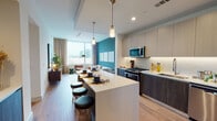 This contemporary urban residence offers a well-appointed single-floor layout designed with modern living in mind. The open-concept kitchen and living area form the heart of the space, where sleek two-tone cabinetry in light gray and dark wood tones pairs with stainless steel appliances, a subway tile backsplash, and a large white quartz waterfall island with bar seating illuminated by pendant lighting. The kitchen flows seamlessly into the living room, which is anchored by a bold teal accent wall and a expansive floor-to-ceiling window that frames sweeping city skyline views and fills the space with natural light. Warm wood-tone flooring runs throughout, tying each area together cohesively. The bedroom continues the city-view experience with a generous window overlooking the urban landscape, complemented by deep navy wall tones that create a calm, sophisticated atmosphere. The primary bathroom is a standout feature, offering a long double-sink vanity with dark wood cabinetry, large-format tile flooring, a glass-enclosed shower, a soaking tub, and direct access to a walk-in closet. A dedicated in-unit laundry area with side-by-side washer and dryer adds everyday convenience. The overall ambiance is modern, curated, and urban.