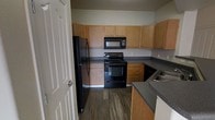 This single-floor residential space offers a practical and comfortable layout designed for everyday living. The dining area is a standout feature, showcasing soft wall-to-wall carpeting underfoot and a curved accent wall that adds a subtle architectural touch. A classic pendant light fixture hangs from the ceiling, providing focused overhead illumination and serving as a simple decorative focal point. Natural light filters in through a nearby window, contributing to a bright and open atmosphere that extends into the adjacent living area.

The kitchen is well-appointed and highly functional, featuring warm honey-toned wood cabinetry that lines the walls from counter to ceiling, offering generous storage throughout. Dark charcoal countertops provide a clean contrast against the cabinetry, while the rich wood-look vinyl plank flooring grounds the space with a contemporary feel. A full suite of black appliances — including a refrigerator, electric range, over-the-range microwave, and dishwasher — delivers a cohesive and complete cooking environment. The u-shaped layout of the kitchen maximizes workspace efficiency and flow. Together, these spaces combine to create a well-organized and welcoming home.