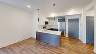 Located in Philadelphia's vibrant South Philadelphia neighborhood, this thoughtfully designed one-bedroom, one-bathroom residence offers a clean, modern aesthetic throughout its efficiently arranged single-floor layout. The open-concept living and kitchen area serves as the heart of the home, bathed in natural light from multiple windows fitted with horizontal blinds. Warm hardwood-style flooring runs seamlessly through the living space, kitchen, and bedroom, tying the unit together with a cohesive, contemporary feel. The kitchen is a standout feature, showcasing a large center island with a crisp white quartz countertop and an integrated stainless steel sink, complemented by sleek dark lower cabinetry and white upper cabinets. A herringbone-patterned tile backsplash adds a stylish touch, while stainless steel appliances—including a range, over-the-range microwave, and full-size refrigerator—complete the modern cooking space. Dramatic cone-shaped pendant lights hang above the island, adding a bold design accent. The bedroom is a bright, serene retreat with recessed lighting, white walls, and a window that allows for natural light. The bathroom features a full soaking tub with elegant large-format marble-look tile surround and a sliding glass shower bar, delivering a clean and polished finish.