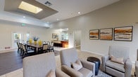 Located in Porter Ranch, Los Angeles, this single-story community clubhouse offers a well-appointed gathering space designed for both social and functional use. The open-concept layout flows seamlessly between key areas, all finished with large-format tile flooring and neutral warm-toned walls that create a bright, welcoming atmosphere throughout.

The central lounge area features plush upholstered armchairs arranged around a patterned area rug, complemented by curated artwork and recessed lighting. A tray ceiling detail adds architectural interest and visual depth to the room. Sliding barn doors lead directly into a dedicated business center, which is lined with built-in shelving and offers a quiet workspace.

The formal dining area accommodates a large dark-wood table with ample seating, set atop hardwood-style flooring and accented with vibrant yellow decorative vases. Tall ceilings and access to exterior doors allow natural light to fill the space. A flat-screen television and whiteboard calendar are also available in the lounge wing, supporting both entertainment and community programming needs.

The fully equipped kitchen features rich wood cabinetry, granite countertops, stainless steel appliances including a refrigerator, range, and microwave, and a generous island with casual seating. Tall indoor plants are positioned throughout the space, adding a natural, refreshing element to the overall interior.
