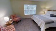 This single-floor residential space offers a warm and inviting atmosphere, highlighted by a thoughtfully appointed bedroom. The bedroom features plush wall-to-wall carpeting that adds softness underfoot and contributes to the room's cozy ambiance. A large window with white plantation-style shutters allows abundant natural light to filter in, offering a pleasant view of the surrounding greenery outdoors. The walls are finished in a soft, neutral tone that enhances the room's bright and airy feel. A comfortable accent chair upholstered in a bold chevron pattern in red and white adds a pop of visual interest and character to the space, complementing the otherwise understated décor. The room provides ample space for a full-sized bed alongside additional seating, making it both functional and comfortable. The overall design reflects a clean, casual aesthetic with practical furnishings and a layout that maximizes usability. The combination of natural light, neutral finishes, and tasteful accent pieces creates a restful retreat that is both welcoming and functional.