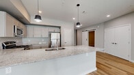 This thoughtfully designed single-floor unit offers a clean, contemporary aesthetic with a neutral color palette and light wood-style flooring that flows seamlessly throughout every room. The open-concept kitchen serves as the centerpiece of the layout, showcasing a large peninsula with granite countertops, an undermount sink, and modern pendant lighting overhead. The kitchen is well-equipped with stainless steel appliances, including a refrigerator, range, and microwave, complemented by soft gray shaker-style cabinetry offering ample storage. A distinctive sliding barn door adds a stylish architectural accent to the living and hallway area, which opens into a spacious, light-filled living room featuring large windows that invite generous natural light. The bedroom features a ceiling fan, recessed lighting, and a closet with built-in wire shelving for organized storage. The bathroom is well-appointed with a granite-topped vanity with shaker cabinetry, a full tub-shower combination, and consistent wood-style flooring that ties the space together. Recessed lighting and clean white trim throughout lend the entire unit a polished, move-in-ready feel. The open floor plan and cohesive design elements create a comfortable and functional living environment.