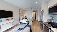 This thoughtfully designed studio with one bathroom offers a smart, open-concept layout that maximizes every square foot. The main living area combines sleeping and lounge spaces in a seamless arrangement, anchored by a sleek four-poster bed with mirrored posts and dressed in crisp white linens accented by teal and navy blue pillows. A comfortable sofa sits just opposite, creating a cozy living zone that flows naturally into the rest of the space. Warm hardwood-style flooring runs throughout, lending continuity and a clean, modern aesthetic. Recessed lighting provides bright, even illumination across the space. The kitchen area is outfitted with stainless steel appliances, including a full-size refrigerator, and features white cabinetry that extends to the ceiling for ample storage. A wall-mounted flat-screen television adds convenience to the living area. Neutral gray-toned walls and white trim contribute to a calm, contemporary palette throughout. A hallway leads to the bathroom and entryway, offering a clear sense of separation between private and shared areas. Overall, this studio presents a well-appointed, efficient living space with modern finishes and a clean, inviting ambiance.