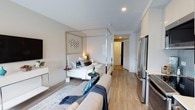 This thoughtfully designed studio with one bathroom offers a smart, open-concept layout that maximizes every square foot. The main living area combines sleeping and lounge spaces in a seamless arrangement, anchored by a sleek four-poster bed with mirrored posts and dressed in crisp white linens accented by teal and navy blue pillows. A comfortable sofa sits just opposite, creating a cozy living zone that flows naturally into the rest of the space. Warm hardwood-style flooring runs throughout, lending continuity and a clean, modern aesthetic. Recessed lighting provides bright, even illumination across the space. The kitchen area is outfitted with stainless steel appliances, including a full-size refrigerator, and features white cabinetry that extends to the ceiling for ample storage. A wall-mounted flat-screen television adds convenience to the living area. Neutral gray-toned walls and white trim contribute to a calm, contemporary palette throughout. A hallway leads to the bathroom and entryway, offering a clear sense of separation between private and shared areas. Overall, this studio presents a well-appointed, efficient living space with modern finishes and a clean, inviting ambiance.
