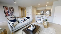 This single-floor residence features an open-concept layout that connects the living area and kitchen into one cohesive, functional space. The living room is anchored by a light grey sofa accented with navy, blush, and white throw pillows, complemented by a dark wood coffee table and a patterned area rug. Recessed lighting illuminates the space with a clean, contemporary feel, while neutral-toned walls and light flooring contribute to an airy, welcoming atmosphere. An accent chair with a cozy white fur throw adds a stylish focal point near the kitchen transition.

The kitchen is defined by warm wood cabinetry, stainless steel appliances including a microwave and range, and a white peninsula counter that doubles as a breakfast bar with wooden barstools. The peninsula provides both additional prep space and a casual dining option. A hallway visible from the main living area leads to additional rooms, suggesting a well-organized floor plan. Overall, the space presents a modern, cohesive design characterized by warm neutral tones, thoughtful furnishings, and an efficient, open layout ideal for everyday living and entertaining.