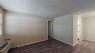 This single-floor unit offers a straightforward, functional layout across several distinct living spaces. The living area features dark hardwood-style flooring and neutral beige walls, complemented by a textured popcorn ceiling. A wall-mounted HVAC unit and a window fitted with horizontal blinds allow for natural light and climate control. A ceiling fan is visible in an adjoining area, which connects directly to the galley-style kitchen.

The kitchen is efficiently arranged in a parallel layout, featuring white cabinetry on both sides, light speckled countertops, a stainless sink, and black appliances including a refrigerator, electric range, and dishwasher. The dark hardwood-style flooring continues throughout. A door at the far end of the kitchen provides additional access.

The bedroom is carpeted in a deep, dark tone and receives natural light through a window with blinds. The room offers adequate wall space and connects to additional areas of the unit.

The bathroom includes a full-size soaking tub with a tiled surround in clean white, a showerhead, and chrome fixtures and accessories. The overall unit presents a neutral, clean palette throughout.