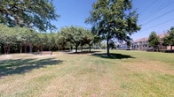 This inviting residential unit is situated directly alongside a serene neighborhood park, offering residents access to open green space shaded by mature trees, park benches, and winding pathways — an ideal setting for relaxation and recreation just steps from the front door.

Inside, the space is characterized by a refined, classic aesthetic. The interior staircase is a notable architectural feature, complete with a polished wood handrail and clean white walls adorned with framed artwork, creating a welcoming transition between levels. Crown molding runs throughout, lending an air of elegance to each room, while warm neutral tones on the walls complement the plush carpeted flooring found across the living areas.

Large windows dressed with floor-length drapes allow generous amounts of natural light to pour in, brightening the interior and creating an airy, open atmosphere. The upper-level landing overlooks the stairwell, offering a sense of vertical depth and spaciousness within the home.

A dedicated sitting or study area provides a comfortable retreat, furnished with a lounge chair and ottoman, a display cabinet with glass doors, and tasteful wall art in calming tones of blue and gold. Track lighting and floor lamps ensure well-lit functionality throughout. Hallways connect the rooms with ease, with closet storage readily accessible. Overall, the space blends comfort, practicality, and thoughtful design in a prime parkside setting.