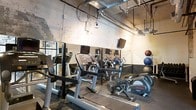 This fitness center offers a comprehensive workout environment designed to accommodate a range of exercise routines. The space features an industrial aesthetic, highlighted by exposed ceiling pipes and ductwork that lend a raw, urban character to the room. Mirrored walls line the space, visually expanding the area and allowing users to monitor their form during workouts. The cardio section is well-stocked with a variety of Life Fitness machines, including treadmills, ellipticals, and stationary bikes, each equipped with individual display screens for entertainment and performance tracking. Wall-mounted televisions are positioned throughout the space, providing additional viewing options. A dedicated functional training area includes medicine balls and other accessories, making the space suitable for dynamic and strength-focused workouts. Overhead lighting, supplemented by pendant fixtures, ensures the gym floor is well-illuminated. The overall ambiance is utilitarian and purposeful, prioritizing function and equipment accessibility. The single-level layout creates an open, navigable floor plan that makes efficient use of the available space.