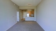 This single-floor residence offers a clean, open-concept layout that seamlessly connects the main living area to the kitchen. The living space is finished with neutral wall-to-wall carpeting and light gray walls, creating a calm and versatile backdrop suited to a variety of furnishing styles. Recessed track lighting in the living area provides focused illumination, while a fluorescent ceiling fixture brightens the kitchen.

The kitchen is positioned at the far end of the open floor plan and features warm wood-toned cabinetry, a stainless steel refrigerator, and a window above the sink that invites natural light into the space. A raised breakfast bar serves as both a functional workspace and a natural divider between the kitchen and the living area, making the layout ideal for entertaining or everyday living.

A hallway extends from the main living space, lined with multiple closet doors offering generous storage. Additional doors along the corridor lead to further rooms, suggesting a well-organized and functional floor plan. The overall ambiance is bright, neutral, and move-in ready, with thoughtful design choices that maximize both utility and openness throughout the single-level layout.