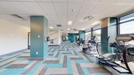 This contemporary fitness center is designed to energize and motivate, combining functional layout with striking visual design. The space is anchored by a bold, typographic wall mural that wraps around key accent walls, featuring fitness-inspired words such as "Energy," "Strength," "Lifestyle," and "Success," setting an uplifting tone throughout. The eye-catching herringbone-patterned carpet in shades of teal, blue, and gray ties the space together and reinforces the vibrant color scheme.

The main exercise area is generously sized and lined with large floor-to-ceiling windows along one side, flooding the room with natural light and offering open outdoor views. This section houses a comprehensive selection of cardio equipment, including ellipticals, treadmills, and stationary bikes, alongside dedicated strength training machines and free weight benches. Teal accent columns punctuate the space, adding architectural interest and visual depth.

A secondary area provides additional room for stretching or targeted training, equipped with benches and supplementary workout equipment. The space also includes a water station near the entrance for added convenience. Recessed panel ceilings with integrated lighting and ceiling fans complete the clean, polished aesthetic. Overall, the fitness center offers a well-organized, inviting environment suited for a full range of workout routines.
