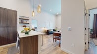 This single-floor residence is designed with an open-concept layout that seamlessly connects the kitchen, dining, and living areas into one cohesive, light-filled space. Soaring ceilings amplify the sense of volume throughout the main living area, while warm-toned pendant lights hang above the kitchen peninsula, adding character and focused illumination.

The kitchen is well-appointed with rich dark wood cabinetry, stainless steel appliances, a white quartz countertop peninsula with bar seating, a subway tile backsplash, and floating shelves that provide both storage and display space. The open living and dining area flows naturally from the kitchen, featuring light hardwood-style flooring that carries throughout the entire residence.

The primary bedroom is generously proportioned, offering natural light through large windows and a distinctive sliding barn door that opens to a walk-in closet. Two full bathrooms are present; one features a tub-shower combination with large-format white tile, while the other includes an elongated vanity with dark wood cabinetry and white countertops. A dedicated laundry room with side-by-side washer and dryer adds everyday convenience. The overall aesthetic is clean, modern, and warm.