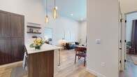 This single-floor residence is designed with an open-concept layout that seamlessly connects the kitchen, dining, and living areas into one cohesive, light-filled space. Soaring ceilings amplify the sense of volume throughout the main living area, while warm-toned pendant lights hang above the kitchen peninsula, adding character and focused illumination.

The kitchen is well-appointed with rich dark wood cabinetry, stainless steel appliances, a white quartz countertop peninsula with bar seating, a subway tile backsplash, and floating shelves that provide both storage and display space. The open living and dining area flows naturally from the kitchen, featuring light hardwood-style flooring that carries throughout the entire residence.

The primary bedroom is generously proportioned, offering natural light through large windows and a distinctive sliding barn door that opens to a walk-in closet. Two full bathrooms are present; one features a tub-shower combination with large-format white tile, while the other includes an elongated vanity with dark wood cabinetry and white countertops. A dedicated laundry room with side-by-side washer and dryer adds everyday convenience. The overall aesthetic is clean, modern, and warm.
