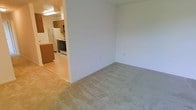 This single-floor residential unit offers a straightforward, functional layout designed for comfortable everyday living. The living area is finished with wall-to-wall carpeting in a neutral tone, creating a warm and understated atmosphere. Smooth, light-colored walls throughout lend a clean, airy feel to the space.

The kitchen is directly accessible from the living area through an open passageway, making the overall floor plan feel connected and practical. The kitchen features tile flooring, wood-toned cabinetry, and includes key appliances such as a refrigerator, a gas range, and a microwave, providing everything needed for daily meal preparation. A countertop peninsula or bar area adds useful workspace and a natural division between the kitchen and the living space.

Overhead lighting in the kitchen ensures the area is well-lit, while the living area benefits from the brightness carried through from the adjoining space. The neutral palette and simple finishes throughout make this a versatile unit, well-suited to a variety of furnishing styles. The seamless transition between the carpeted living space and the tiled kitchen highlights the open, cohesive nature of the floor plan.