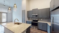 This single-floor unit showcases a thoughtfully designed open layout with soaring ceilings that lend an airy, expansive feel throughout the living areas. Rich wood-look plank flooring anchors the main living space, which flows seamlessly into a well-appointed kitchen. The kitchen is a standout feature, boasting gray shaker-style cabinetry extending to the ceiling, light granite countertops, a gray tile backsplash, and a full suite of stainless steel appliances including a range, over-the-range microwave, and a French door refrigerator. A central island with an undermount sink and pendant lighting provides additional prep space and casual dining. The living area benefits from generous natural light through French doors fitted with blinds, complemented by a transom window above that further amplifies the lofty ceiling height. Bedrooms feature plush carpeting, neutral walls, and large windows that invite natural light. The primary bedroom includes access to a spacious walk-in closet outfitted with multiple hanging rods, built-in shelving, and generous storage capacity. The bathroom features a dark-framed vanity mirror, granite countertop, undermount sink, and coordinating gray cabinetry, maintaining the unit's cohesive modern aesthetic throughout.