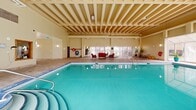 Located at 160 Chapel St in Ottawa's Sandy Hill neighbourhood, this indoor aquatic facility offers a well-appointed pool area designed for both leisure and fitness. The centerpiece is a generously sized indoor lap pool with vivid turquoise water, tiled in classic blue mosaic and equipped with stainless steel entry rails and shallow-end steps, clearly marked with depth indicators and posted health regulations for user safety.

The space is defined by its striking coffered ceiling with exposed structural beams, lending the room an open and airy industrial-meets-institutional character. Large floor-to-ceiling windows and a glass-enclosed patio wall flood the interior with natural light, creating a bright and inviting atmosphere throughout the day. The patio area adjacent to the pool provides a comfortable seating arrangement with red upholstered chairs, ideal for relaxing between swims or supervising activity in the water.

The surrounding deck is finished with neutral stone-toned tile, offering a clean and practical walking surface. Wall-mounted sconces complement the ambient lighting, while safety equipment including a life ring and fire hose cabinet are visibly positioned throughout. Ancillary areas include a tiled transition corridor with access to change facilities, complete with proper ventilation and compliance signage, ensuring a fully functional and regulation-compliant aquatic environment.