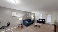 This single-floor residence offers a functional and comfortable layout across three bedrooms, three bathrooms, a living room, a dedicated office, and a kitchen. The living room features neutral gray walls, plush carpeting, and a window that allows natural light to fill the space, creating a welcoming atmosphere. The kitchen is equipped with white cabinetry, dark granite-style countertops, white appliances, and hardwood-style flooring, and opens directly toward the living area for a connected feel. Hallways with carpeted floors connect the bedrooms and bathrooms, maintaining a cohesive flow throughout the unit. The bedrooms are carpeted and include ceiling fans, closets, and windows with blinds for natural light and privacy. The dedicated office space features a ceiling fan, a window, and carpeted flooring, making it a practical workspace. The bathrooms feature vanities with dark countertops and white cabinetry. The building exterior is clad in teal siding with brick accents, and ground-floor units have direct outdoor access to a shared grassy area. Upper levels feature balconies with metal railings. Three closets provide ample storage throughout the residence.