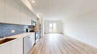 This single-floor unit offers a thoughtfully designed layout that maximizes its compact footprint. Light hardwood flooring runs continuously throughout, creating a cohesive and airy feel across all areas. The open-plan kitchen and living area forms the heart of the unit, where natural light streams in through a well-positioned window offering outdoor views of the surrounding neighborhood. The living area provides a clean, versatile space with crisp white walls.

The kitchen is outfitted with a full suite of stainless steel appliances, including a refrigerator, range, over-the-range microwave, and dishwasher. White flat-panel cabinetry extends to the ceiling, maximizing storage, while a distinctive blue subway tile backsplash adds a bold design accent against the light countertops. A single-basin sink with a modern faucet completes the functional workspace.

A hallway connects the main living area to the bathroom, which features a full soaking tub with shower, tiled surround in white, and brushed nickel fixtures. Recessed lighting and white walls maintain the unit's consistently clean, modern aesthetic. Throughout the space, a neutral white palette, recessed lighting, and light wood tones combine to create a bright, welcoming atmosphere.