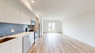This single-floor unit offers a thoughtfully designed layout that maximizes its compact footprint. Light hardwood flooring runs continuously throughout, creating a cohesive and airy feel across all areas. The open-plan kitchen and living area forms the heart of the unit, where natural light streams in through a well-positioned window offering outdoor views of the surrounding neighborhood. The living area provides a clean, versatile space with crisp white walls.

The kitchen is outfitted with a full suite of stainless steel appliances, including a refrigerator, range, over-the-range microwave, and dishwasher. White flat-panel cabinetry extends to the ceiling, maximizing storage, while a distinctive blue subway tile backsplash adds a bold design accent against the light countertops. A single-basin sink with a modern faucet completes the functional workspace.

A hallway connects the main living area to the bathroom, which features a full soaking tub with shower, tiled surround in white, and brushed nickel fixtures. Recessed lighting and white walls maintain the unit's consistently clean, modern aesthetic. Throughout the space, a neutral white palette, recessed lighting, and light wood tones combine to create a bright, welcoming atmosphere.