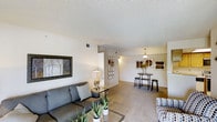 This single-floor residence offers a welcoming and well-appointed open-concept layout that seamlessly connects the living and dining areas. The living room is anchored by a comfortable sectional sofa in a cool grey tone, complemented by a glass coffee table, a patterned accent chair, and lush indoor plants that add a fresh, natural element to the space. A framed landscape artwork adorns the wall, adding a touch of character to the room. Soft, neutral-toned carpeting runs throughout the main living area, contributing to a warm and inviting ambiance. Overhead, a textured ceiling with integrated ventilation adds a classic residential feel. Decorative circular mirror wall art serves as a tasteful focal point along the transitional wall leading toward the dining area. The dining space features a sleek bar-height table with matching stools beneath a pendant light, creating an intimate setting for casual dining or entertaining. Framed artwork continues into the dining area, maintaining a cohesive and curated aesthetic. The overall design palette of whites, greys, and warm neutrals gives the space a clean, modern, and comfortable character throughout.