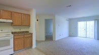 This single-floor space presents a straightforward, functional layout that transitions seamlessly from a kitchen area into an open living space. The kitchen section, positioned along one side, features warm honey-toned wood cabinetry with a granite-look countertop, providing ample storage and workspace. The flooring in the kitchen area consists of light-colored tile, creating a natural visual boundary as it gives way to the wall-to-wall carpeting that covers the expansive main living area.

The living space is finished with smooth, white walls and a clean white ceiling, giving the area a bright and neutral ambiance. Overhead HVAC vents are integrated into the ceiling, while baseboard vents along the walls ensure consistent climate control throughout. Multiple electrical outlets are distributed across the walls, offering practical utility for various furniture arrangements.

A doorway at the far end of the living area leads to an additional room, visible through the opening and similarly carpeted, suggesting continuity in the overall design. The space's open configuration and neutral palette provide a versatile foundation suitable for a variety of uses and personal styling preferences.