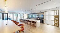This single-floor amenity space is designed with a refined, contemporary aesthetic that seamlessly blends social and functional zones. The open-plan layout is anchored by a striking, elongated white stone kitchen island with bar seating, complemented by sleek dark cabinetry, a gas cooktop, and a textured backsplash beneath dramatic pendant lighting. A mirrored wall behind the kitchen counter amplifies depth and light throughout the space.

The lounge areas feature plush white sofas accented with rich burnt-orange and patterned throw pillows, arranged atop bold geometric rugs in navy and red. Elegant table lamps, gold-toned fixtures, and curated wall art contribute to the upscale atmosphere. Ornate gold lattice partitions serve as decorative dividers, adding architectural interest between zones.

Floor-to-ceiling windows and glass sliding doors line multiple walls, flooding the interior with natural light and providing direct views of an outdoor terrace. Light hardwood flooring flows through the kitchen and dining areas, while carpeted lounge sections define intimate seating arrangements. An adjacent grand lobby space, visible through glass walls, features a dramatic spiral staircase, a suspended fireplace, and double-height ceilings, enhancing the overall sense of grandeur.