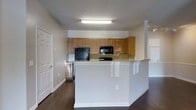 This single-floor space centers around an open-concept kitchen designed for both functionality and accessibility. The kitchen is anchored by a large, white peninsula that extends into the living area, offering ample counter space and serving as a natural divider between the cooking and living zones. The peninsula's clean white finish contrasts with the warm honey-toned wood cabinetry that lines the kitchen walls, providing generous upper storage across multiple cabinet banks. Black appliances, including a refrigerator, microwave, and cooktop, are integrated seamlessly into the cabinetry, lending a cohesive and practical aesthetic. A light-toned countertop runs the length of the kitchen, complementing the overall neutral color palette of beige and white walls. Dark hardwood flooring spans the visible areas of the space, adding warmth and visual contrast against the lighter finishes. Overhead, a flush fluorescent light fixture provides direct illumination to the kitchen workspace. A paneled white door is visible to the left, suggesting access to an additional room. Decorative column details near the peninsula add a subtle architectural element to the open layout.