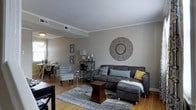 This two-story home offers a thoughtfully designed interior that balances comfort and style across its multiple levels. The main living area serves as the heart of the home, showcasing warm hardwood flooring that extends throughout the open-concept space. Crown molding frames the ceilings, adding a refined architectural detail that elevates the overall aesthetic.

The living room is generously proportioned, providing ample space for relaxed seating arrangements. Neutral gray walls create a versatile backdrop that complements both the natural wood tones and the carefully chosen furnishings. The space flows naturally into a dining area positioned near a window, allowing natural light to filter in and brighten the interior.

A decorative sunburst mirror mounted on the main wall serves as a focal point in the living area, adding texture and visual interest. The open layout promotes a seamless connection between the living and dining zones, making the space well-suited for both everyday living and entertaining. Vaulted ceiling detailing and visible ductwork contribute to the home's functional yet stylish character. The two-story configuration provides additional living space above, offering a distinct separation between common areas and private quarters.