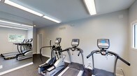 Located at 1100 Audubon Parc Dr in Cary, NC, this fitness center offers a dedicated space for residents to maintain an active lifestyle. The single-floor facility is designed with functionality and comfort in mind, featuring a clean, open layout with light gray walls and dark, durable flooring suited for high-traffic exercise use. Overhead fluorescent lighting runs the length of the ceiling, ensuring the space is evenly and brightly illuminated throughout the day. The room is equipped with a lineup of True-brand cardio machines, including treadmills and elliptical trainers, positioned to allow comfortable use and easy movement between equipment. A full-length mirrored wall along one side of the room enhances the sense of openness while allowing users to monitor their form during workouts. Climate control is provided via a ceiling vent, keeping the environment comfortable year-round. A private door leads to an adjoining room, suggesting additional facilities or storage on-site. The overall ambiance is modern, clean, and purpose-built — a practical and well-appointed fitness amenity designed to serve the needs of the surrounding community.
