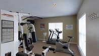 This single-floor fitness room offers a practical and functional space for exercise, designed to accommodate a variety of workout routines. The room features a multi-station cable weight machine positioned along one wall, providing options for strength training exercises. A treadmill and a True-brand stationary bike are also present, catering to cardiovascular workouts. The neutral tan walls and carpeted flooring create a modest, straightforward environment. Recessed lighting in the textured ceiling provides adequate illumination throughout the space. A window fitted with white horizontal blinds allows for natural light while maintaining privacy. A standing fan is available for additional ventilation and comfort during workouts. Informational posters, including a target heart rate chart, are displayed on the walls, offering guidance to users. A posted notice indicates age restrictions for use of the facility, suggesting this is a shared amenity space. Overall, the room is efficiently laid out to maximize the available floor area while housing essential fitness equipment for general use.