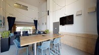 Located at 712 Tucker St in Raleigh, NC, this single-floor amenity space seamlessly blends indoor comfort with open-air living. The area is defined by dramatically tall ceilings that amplify the sense of volume and openness throughout. Warm string globe lights are strung overhead, creating an inviting and social atmosphere during any time of day.

The space is divided into distinct functional zones. A lounge area features plush wicker sofas and armchairs with gray cushions, teal-patterned throw pillows, and a glass-top coffee table, all arranged atop a vibrant geometric area rug. Colorful abstract artwork adorns the walls, adding a coastal-inspired aesthetic. Stone masonry wainscoting lines the lower walls, adding a grounded, textural contrast to the bright painted upper walls and white panel molding details.

Multiple bar-height tables paired with modern perforated-back stools serve as gathering and dining zones, each positioned near wall-mounted flat-screen televisions. Navy blue floor-to-ceiling curtains frame open archways that look out directly onto a landscaped courtyard and pool area, flooding the space with natural light and outdoor views. Potted snake plants provide natural greenery throughout. The polished aggregate flooring ties the entire space together, making it a versatile and well-appointed communal destination.