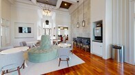 Located at 101 Breakers Dr in Myrtle Beach, SC, this single-floor community clubhouse offers an elegantly designed gathering space that blends coastal charm with refined interior details. The main lounge area immediately impresses with soaring double-height ceilings, floor-to-ceiling windows, and an abundance of natural light that floods the rich hardwood flooring throughout. Large multi-pane windows frame views of the surrounding outdoor pool area and lush landscaping, connecting the interior seamlessly to the outdoors.

The central social space is anchored by a distinctive circular tufted mint-green sofa, complemented by an assortment of white accent tables and stylish side chairs. Overhead, a striking geometric chandelier and elegant pendant lights add a sophisticated ambiance to the coffered ceiling above. A dedicated bar-height seating area lines one wall beneath a decorative wallpapered accent, providing a casual space for residents to gather.

A coffee and refreshment station is conveniently built into the wall, offering easy access for daily use. The entryway features polished concrete flooring in a muted green tone, arched doorways, and wall sconces that create a warm, welcoming arrival experience. An adjacent lounge nook with striped accent walls, bench seating, and pedestal tables provides a quieter space for conversation or work. The overall design balances coastal elegance with functional, community-oriented living.