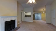 This single-floor residence offers a thoughtfully designed open layout that maximizes both comfort and functionality. The living area is an immediate focal point, distinguished by soaring vaulted ceilings that create an expansive, airy atmosphere. A ceiling fan with a light fixture is centrally positioned to complement the generous vertical space. A classic white-mantled fireplace with brick detailing anchors one wall, adding warmth and a traditional architectural character to the room. Plush, neutral-toned carpeting spans the living space, contributing to the overall sense of comfort and continuity throughout. A pass-through window connects the living area to the kitchen, which features white cabinetry, a dark countertop, and a practical layout designed for efficient use of space. This open connection between the two areas promotes an easy flow ideal for both everyday living and entertaining. Natural light fills the space through a large sliding glass door dressed with vertical blinds, offering convenient access to the outdoors. A decorative chandelier is visible in an adjacent area, hinting at a defined dining space nearby. The warm, beige color palette used throughout unifies each area cohesively, resulting in a welcoming and well-balanced interior.