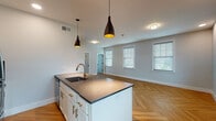 Located at Johnson Mill Blvd in Johnson, Arkansas, this single-floor residence offers a thoughtfully designed open-concept layout with contemporary finishes throughout. The striking herringbone-patterned hardwood flooring is a defining feature, flowing seamlessly through every room and lending warmth and visual interest to the entire space.

The kitchen serves as a central hub, anchored by a large island with a dark countertop, an undermount sink, and a brushed gold faucet. White shaker-style cabinetry with gold hardware complements the stainless steel appliances, which include a refrigerator, microwave, and range. Sleek black pendant lights with gold interiors hang above the island, adding a modern yet warm ambiance.

The open living and dining area is bathed in natural light from multiple windows fitted with white blinds, creating a bright and airy atmosphere. The neutral gray walls provide a clean, versatile backdrop throughout.

The bedrooms are well-proportioned, each featuring herringbone floors, natural light, and ceiling fans for comfort. A dedicated laundry closet houses a stacked washer and dryer unit, maximizing convenience within a compact footprint. The bathroom features white cabinetry with gold hardware, a dark stone countertop, an undermount sink, and a matte black faucet, maintaining the cohesive modern aesthetic found throughout the home.