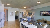 This single-floor studio showcases a thoughtfully designed open-concept layout that maximizes functionality and visual flow. The exposed concrete ceiling serves as a defining architectural feature, lending an industrial character balanced by warm hardwood flooring that runs throughout the main living area. The sleeping area is centrally anchored by a generously sized bed dressed in blue and yellow tones, flanked by matching nightstands with wall-mounted sconce lighting and a framed piece of artwork above the headboard. A mirrored closet system provides practical storage while reflecting light across the space. The dining area features a sleek white bar-height table paired with modern counter stools, seamlessly transitioning into the living area beyond. Flush-mount ceiling fixtures provide ambient lighting throughout. A separate bathroom is accessible through a doorway, visible in the background, with a vanity area partially in view. The muted blue-grey accent wall in the sleeping zone adds depth and contrast to the otherwise neutral palette, while the overall design balances urban, industrial elements with warm, residential comfort.