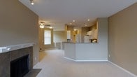 This single-floor residence offers an open-concept layout that seamlessly connects the living and kitchen areas, creating a spacious and functional environment. The living room is anchored by a fireplace, adding a warm focal point to the space, while light-toned carpeting extends throughout, contributing to the room's inviting atmosphere. A ceiling fan with integrated lighting provides both comfort and illumination. Recessed lighting is installed across the ceiling, ensuring even, practical light distribution throughout the open area.

The kitchen is positioned at the far end of the space and features white cabinetry, a refrigerator, and a range with a microwave above. A peninsula with a dark countertop serves as a natural divider between the kitchen and living area, offering additional workspace and potential seating. The cabinetry extends to a second tier, maximizing vertical storage. Neutral taupe walls tie the rooms together cohesively, while a window in the living area allows natural light to filter in. The layout and design elements combine to create a practical, well-connected living environment.