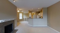 This single-floor residence offers an open-concept layout that seamlessly connects the living and kitchen areas, creating a spacious and functional environment. The living room is anchored by a fireplace, adding a warm focal point to the space, while light-toned carpeting extends throughout, contributing to the room's inviting atmosphere. A ceiling fan with integrated lighting provides both comfort and illumination. Recessed lighting is installed across the ceiling, ensuring even, practical light distribution throughout the open area.

The kitchen is positioned at the far end of the space and features white cabinetry, a refrigerator, and a range with a microwave above. A peninsula with a dark countertop serves as a natural divider between the kitchen and living area, offering additional workspace and potential seating. The cabinetry extends to a second tier, maximizing vertical storage. Neutral taupe walls tie the rooms together cohesively, while a window in the living area allows natural light to filter in. The layout and design elements combine to create a practical, well-connected living environment.