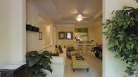 This single-floor residence offers a thoughtfully designed open-concept layout that seamlessly connects the living area to the kitchen, creating a spacious and cohesive flow throughout. The living room is a welcoming focal point, centered around plush white sofas, patterned accent chairs, and an upholstered ottoman that doubles as a coffee table. A tray ceiling with a ceiling fan adds architectural interest overhead, while soft, neutral wall tones and warm lighting contribute to an inviting and comfortable ambiance. Lush indoor plants are positioned throughout, bringing a touch of greenery that enlivens the space. The kitchen is visible from the living area and features white cabinetry, a full-size refrigerator, and a raised breakfast bar with elegant bar stools, making it ideal for casual dining and entertaining. An accent wall in a rich green hue adds a bold contrast to the otherwise light and airy color palette. Framed artwork on the walls adds a curated, homey feel. The consistent use of light flooring and neutral furnishings throughout the space enhances the sense of openness, making this residence a well-balanced blend of comfort, style, and functionality.