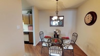This single-floor residential space offers a welcoming and functional layout designed for comfortable living. The dining area serves as a charming focal point, featuring warm hardwood-style flooring that adds richness and continuity to the space. A round glass-top dining table paired with ornate wrought-iron chairs creates an elegant yet inviting setting, illuminated by a classic chandelier that hangs gracefully from the ceiling. A decorative framed mirror mounted on the neutral-toned wall further enhances the sense of light and openness in the room. Adjacent to the dining area, the kitchen is efficiently arranged with dark wood cabinetry, light countertops, and essential appliances, including a dishwasher, making it practical for everyday use. A textured ceiling adds subtle character throughout the space. The overall ambiance is warm and homey, with a cohesive color palette of neutral walls and rich wood tones that tie the rooms together seamlessly. The open sightline between the kitchen and dining area promotes a natural flow, making the space feel connected and easy to navigate.
