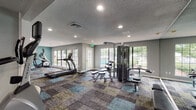 This single-floor amenity facility at 5900 Old Hickory Blvd in Nashville, Tennessee offers residents a well-appointed fitness center and a dedicated laundry room. The fitness center is a generously sized, open workout space finished with stylish multi-toned carpet tiles in shades of teal, gray, and gold. The room is bathed in natural light through multiple large windows and glass-panel doors that offer views of the surrounding outdoor areas, including a pool. A full-length mirror wall visually expands the space and supports a variety of training routines. Equipment includes treadmills, an elliptical, a multi-station cable machine, adjustable benches, and additional strength-training apparatus. The walls are painted in a combination of cool gray and a bold teal accent, giving the space a modern, energetic feel. Recessed lighting and a textured ceiling complete the interior. Adjacent to the fitness area, the laundry room provides a clean, functional environment with rows of front-loading washers and dryers, smart-card-enabled machines, and wood-look plank flooring. The room is well-lit and neatly organized, with posted instructional signage for user convenience. Together, these amenities reflect a thoughtfully designed, practical community facility.