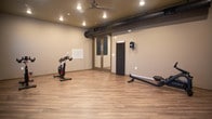 Located at 548 Newtown Rd in Virginia Beach, Virginia, this single-floor fitness studio is purpose-built for focused, equipment-based workouts. The space features warm hardwood-style plank flooring throughout, providing a clean and durable surface well-suited for a variety of exercise activities. The interior is finished with neutral gray walls and a dark ceiling accented by recessed can lighting and ceiling-mounted track lights, creating a modern, energizing atmosphere. Exposed black ventilation ductwork runs along one wall, adding an industrial character to the design. Multiple stationary cycling bikes are positioned throughout the open floor plan, complemented by a rowing machine, offering a well-rounded cardio training environment. A large flat-screen television is wall-mounted, ideal for streaming workout classes or fitness content during sessions. Natural light filters in through several windows along one wall, providing views of the surrounding area and keeping the space feeling open and connected to the outdoors. Glass-panel doors offer a sleek entryway and visual transparency between areas. The layout is open and unobstructed, allowing for flexible equipment arrangements or group fitness classes. Overall, the studio delivers a clean, functional, and motivating environment for cardiovascular training.