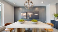 This well-appointed meeting room delivers a refined yet characterful atmosphere through a thoughtfully curated interior design. The focal point of the space is a dramatic dark slate-gray slatted accent wall, against which a triptych of large-format Western longhorn photographs is displayed, lending the room a bold and memorable identity. A substantial marble-topped conference table anchors the center of the room, surrounded by cognac leather barrel chairs with gold-toned metal frames — a combination that strikes a balance between sophistication and warmth. Overhead, a rustic orb chandelier with rope detailing and candelabra-style lighting adds a layer of organic texture to the otherwise contemporary setting. The room is carpeted with a neutral, multi-toned loop pile rug that complements the earthy palette throughout. A built-in credenza with navy blue cabinetry and a light stone countertop lines one wall, topped with a wall-mounted flat-screen display — making the space well-suited for presentations or collaborative meetings. Large windows allow natural light to filter in from an adjacent outdoor area, while interior windows and pass-throughs connect the room to the broader facility. Recessed ceiling lighting provides consistent ambient illumination, completing a space that is both functional and visually engaging.