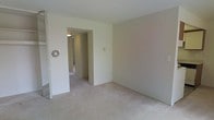 This single-floor space presents a clean, understated interior defined by neutral beige walls and wall-to-wall carpeting that runs consistently throughout the visible areas. The main room is open and simply appointed, offering a blank canvas well-suited for a variety of furniture arrangements. A rectangular doorway opening leads into a hallway, which connects to additional rooms beyond, suggesting a functional and straightforward floor plan. A circular ventilation grille is mounted on the wall near the entry, contributing to the unit's climate control system. The hallway, illuminated by a ceiling-mounted light fixture, provides a glimpse of further rooms, including what appears to be a door at the far end. The walls are smooth and uniformly painted, complementing the light-toned carpet underfoot. Overall, the space maintains a modest, practical ambiance with consistent finishes throughout, offering a neutral foundation that can be easily personalized.