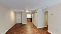 This single-floor residence offers a clean, open layout finished with rich hardwood-style flooring that runs throughout the main living areas, lending warmth and continuity to the space. The entry area is bright and welcoming, featuring a classic six-panel front door with a mail slot, setting a tidy and functional tone from the moment you arrive. Track lighting overhead provides well-distributed illumination across the open corridor and living zone. A standout feature of the main hallway is a set of full-height, double-panel closet doors, offering generous built-in storage that keeps the living areas clean and uncluttered. The kitchen is visible from the main living space and showcases warm wood cabinetry, a dark countertop, and a stainless steel sink positioned beneath a window that allows natural light to brighten the workspace. Dark appliances complement the cabinetry, creating a cohesive and practical kitchen aesthetic. The neutral, light-toned walls throughout the space enhance the sense of openness and provide a versatile backdrop for a variety of interior styles. The overall ambiance is functional and move-in ready, with a layout that encourages easy flow between the entry, living, and kitchen areas.