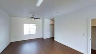 This well-maintained two-floor space offers a thoughtfully designed interior that balances simplicity with functionality. The living area immediately draws attention with its rich, dark hardwood-style flooring that runs throughout, providing a warm contrast against the crisp white walls and bright white ceilings. A modern ceiling fan with integrated lighting anchors the main living space, ensuring year-round comfort and airflow. Natural light filters in through a window fitted with clean white blinds, lending the room an airy, open feel. The neutral color palette throughout creates a versatile backdrop, making the space easy to personalize. The layout flows naturally, with clear sightlines from the living area toward the entryway, which features a solid front door and a dedicated closet — ideal for storage needs. Recessed and accent lighting complements the natural light, ensuring the space remains bright at any hour. The clean, minimalist aesthetic is consistent across the interior, reflecting a modern design sensibility. Spanning two floors, the space offers ample room for both living and private quarters, making it a practical and inviting environment for a variety of lifestyles.